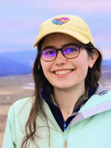Young woman with brown hair and yellow cap in front of grassland and mountains.