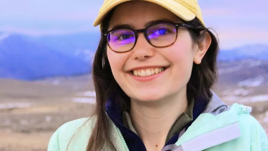 Young woman with brown hair and yellow cap in front of grassland and mountains.